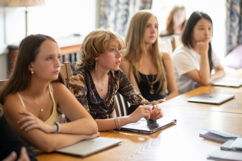 Students sit a table listening to a lecture some raising their hands