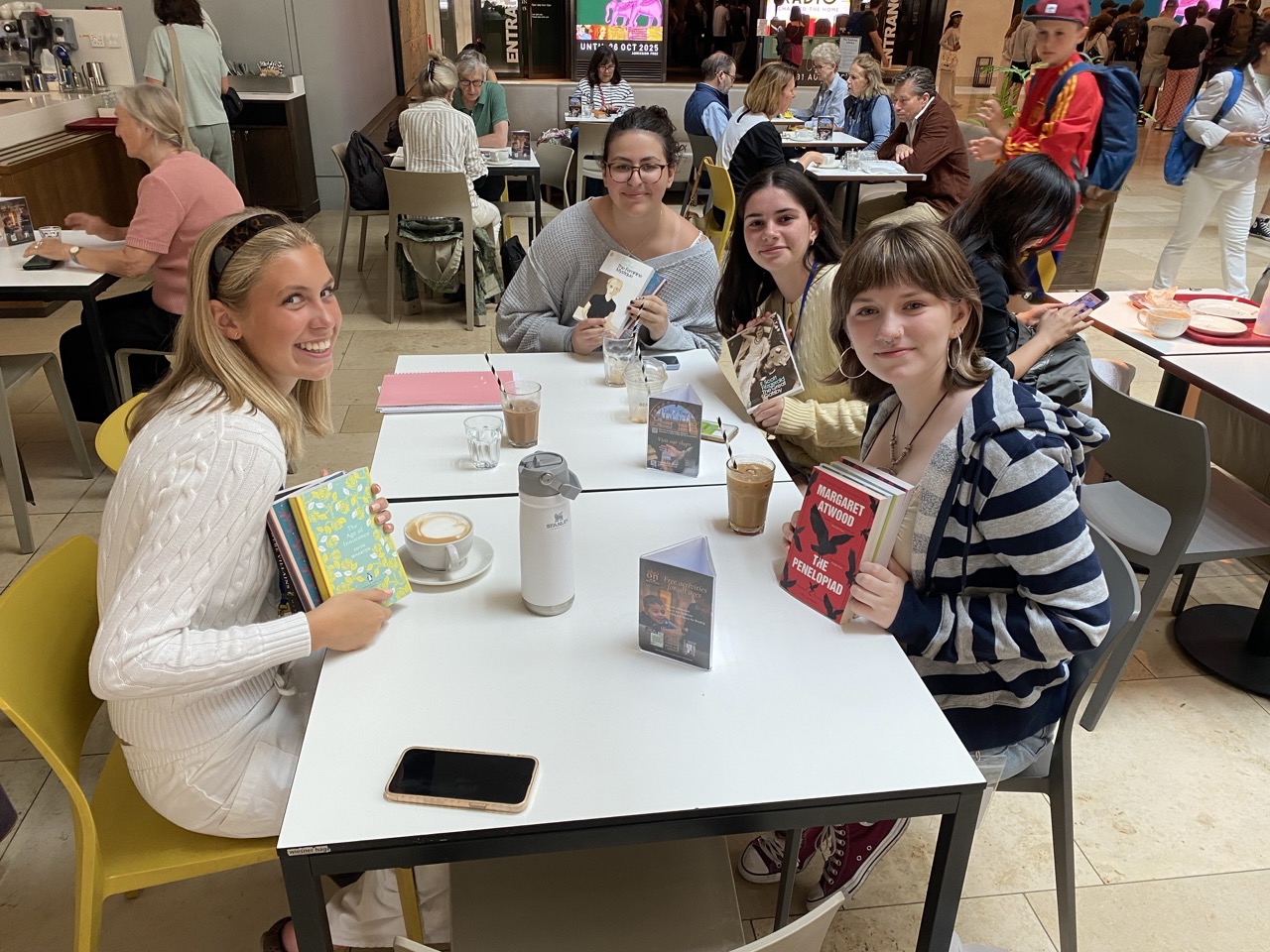 Four students sit at a cafe table and hold up books