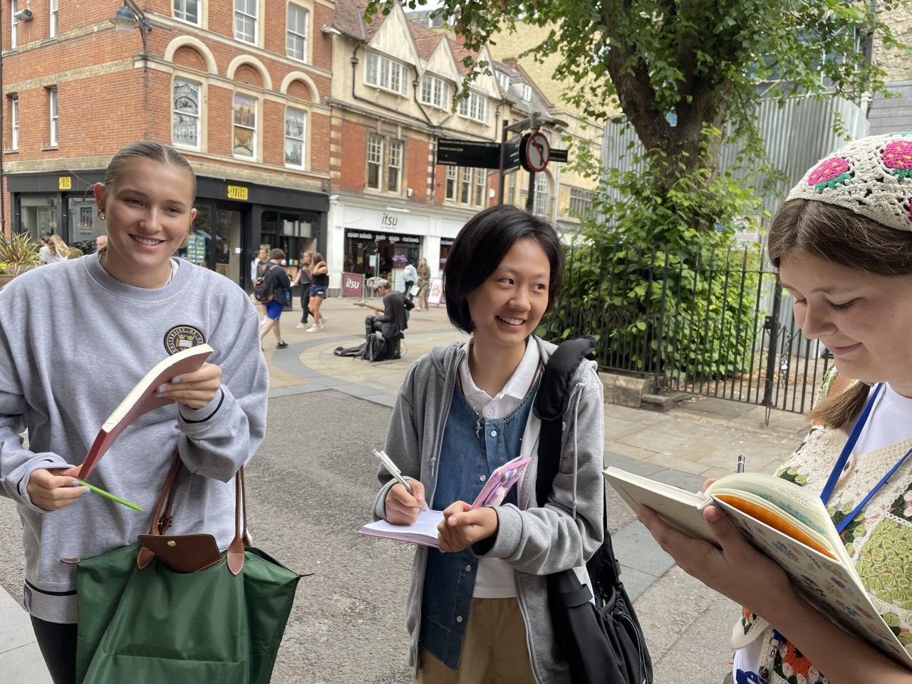 Three smiling students stand on streets on Oxford writing in notebooks
